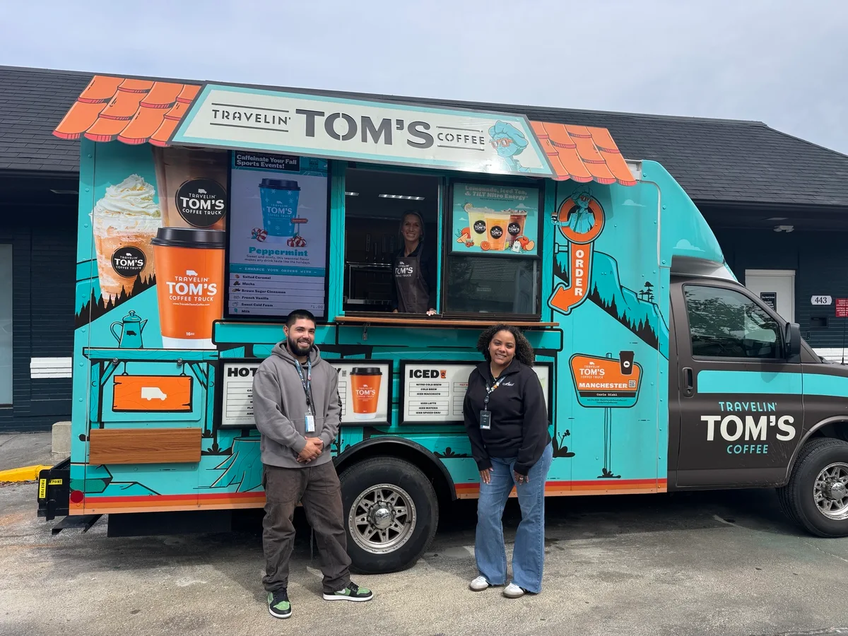 Two people wearing lanyards stand in front of a turquoise Travelin' Tom's Coffee truck in a parking lot.